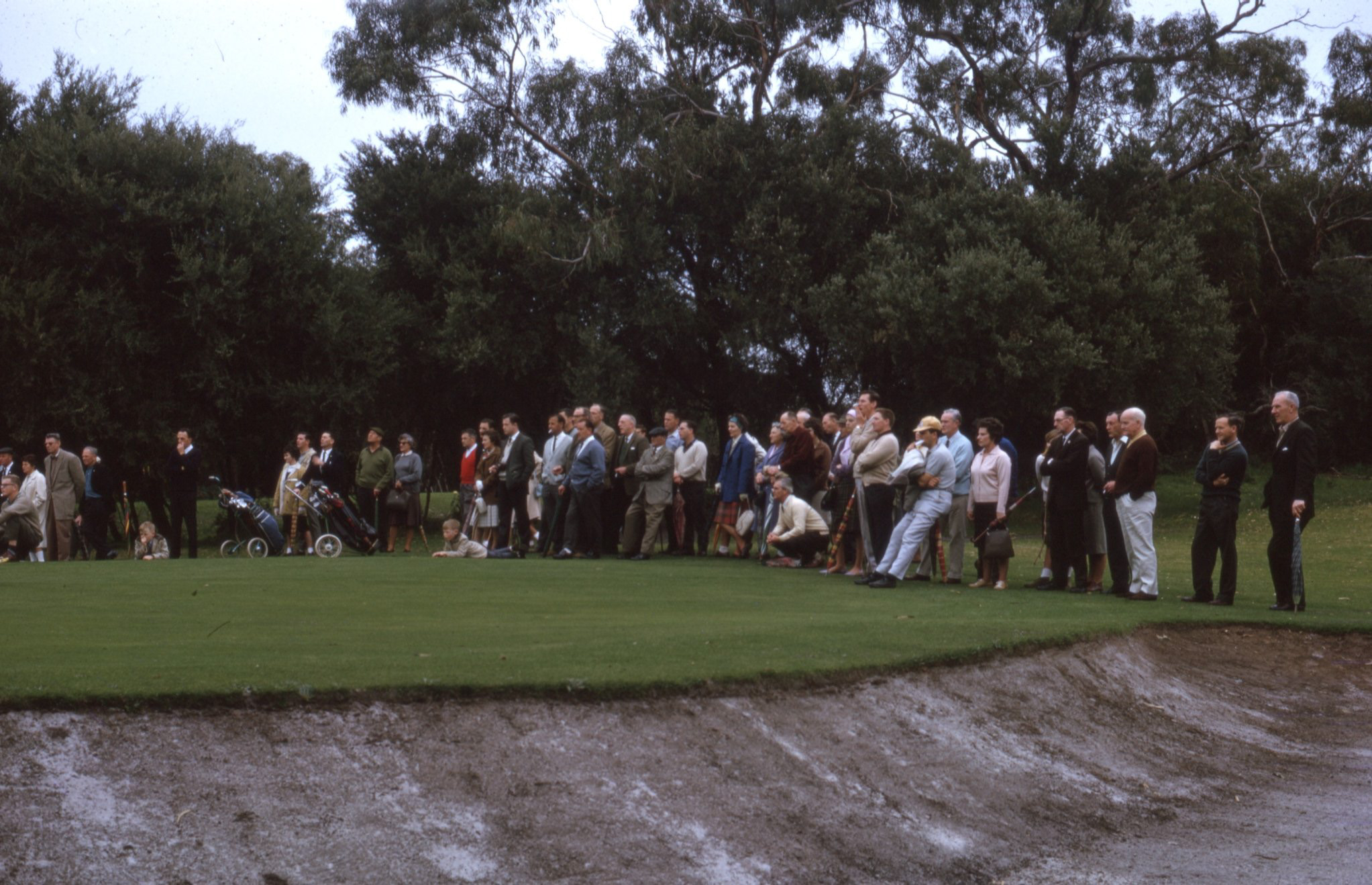 A crowd of spectators in 1950s golfing attire watching a tournament from the edge of the green