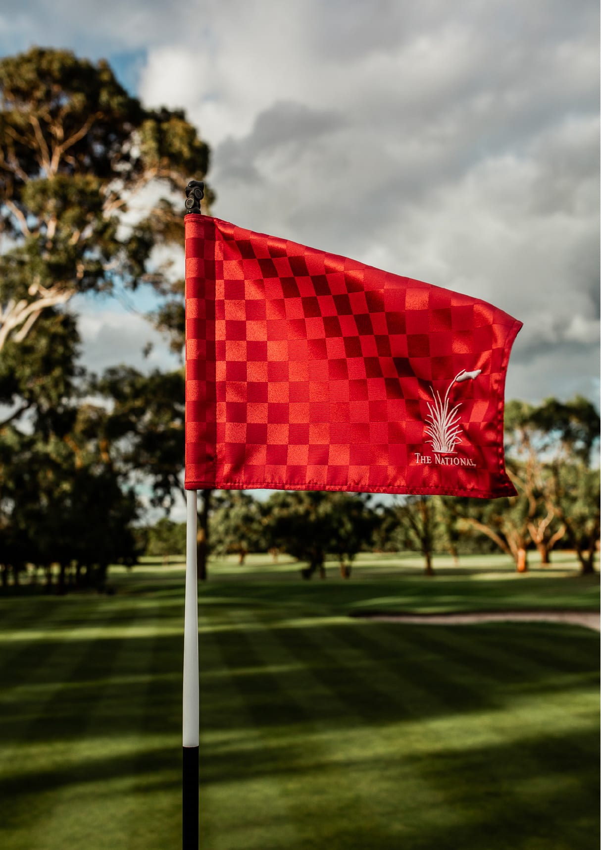 The National Golf Club's red chequered flag on a flagstick above a Long Island green, with gum trees beyond